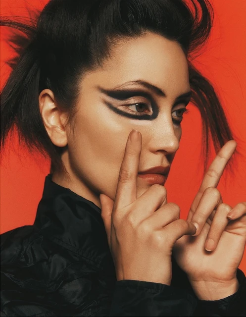 Woman with dramatic black eyeshadow and neutral lips, touches her face in front of a plain coral backdrop, wearing black jacket.