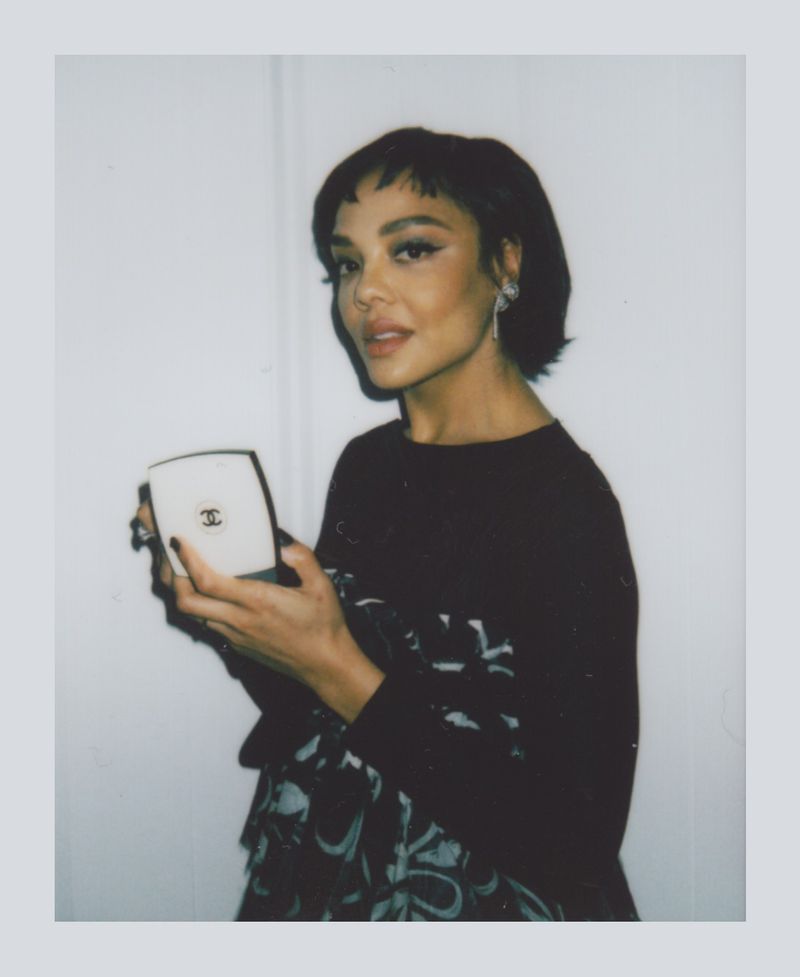 A woman with short dark hair holds a compact mirror in a portrait shot against a white background, framed by a polaroid border.