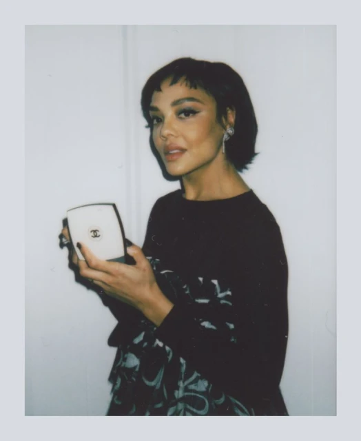 A woman with short dark hair holds a compact mirror in a portrait shot against a white background, framed by a polaroid border.
