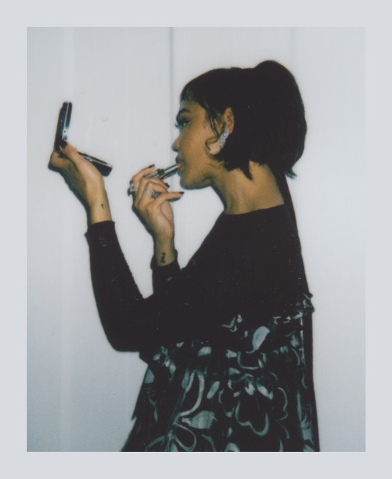 Young woman applies lipstick in a photo studio, using a compact mirror. She wears a floral top, and there is soft studio lighting.