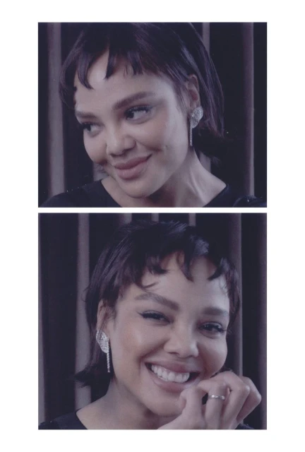 Close-up portrait of a smiling woman with short brown hair and diamond earrings against a dark grey curtain backdrop in soft lighting.