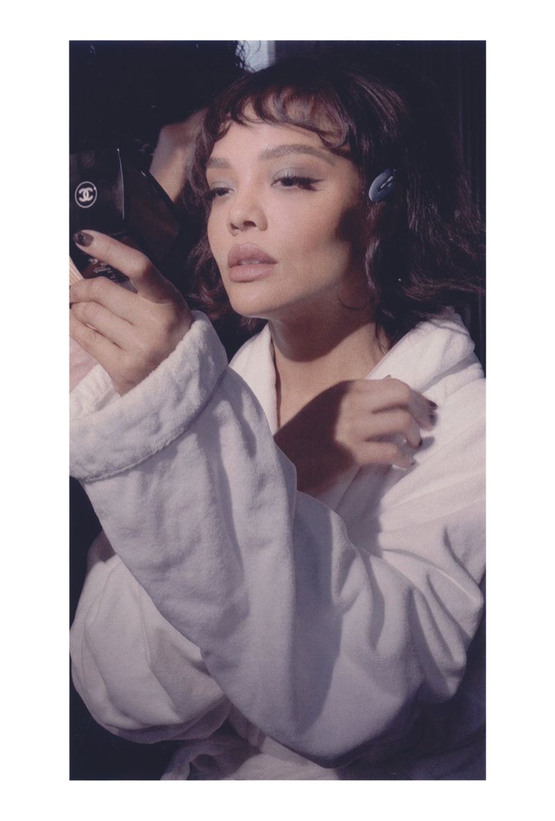 Adult woman in white robe poses indoors holding a makeup compact; eyeshadow, hair clips, and dark nail polish visible in dim light.