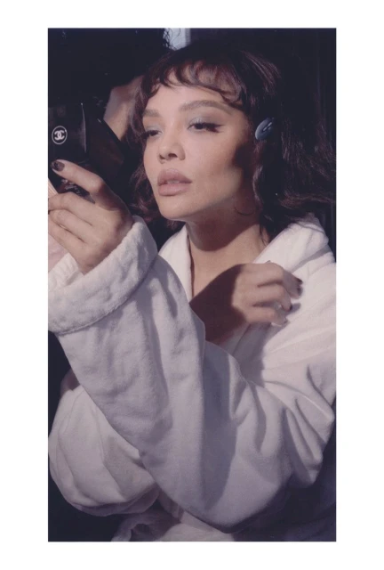 Adult woman in white robe poses indoors holding a makeup compact; eyeshadow, hair clips, and dark nail polish visible in dim light.