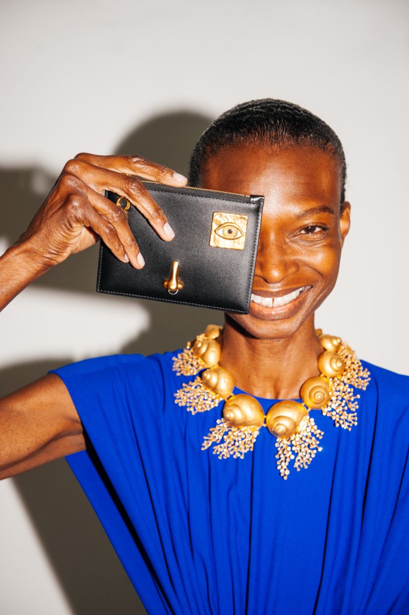 A dark-skinned woman smiles, holding a black clutch with a gold accent, and wearing a large gold seashell necklace and a blue dress.