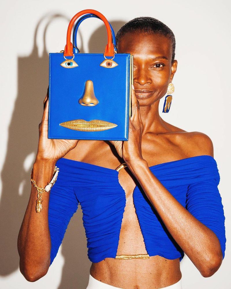 Woman in blue top poses with blue handbag featuring a face design. The handbag has a nose, eyes, and lips. White background.