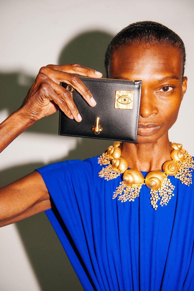 An adult female model poses indoors wearing a blue dress and jewelry, holding a black wallet with gold decorative elements.