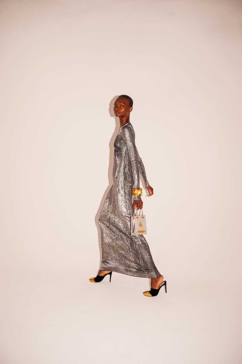A black female model walking in a shiny silver gown in a studio with a bracelet and small purse, against a white background.