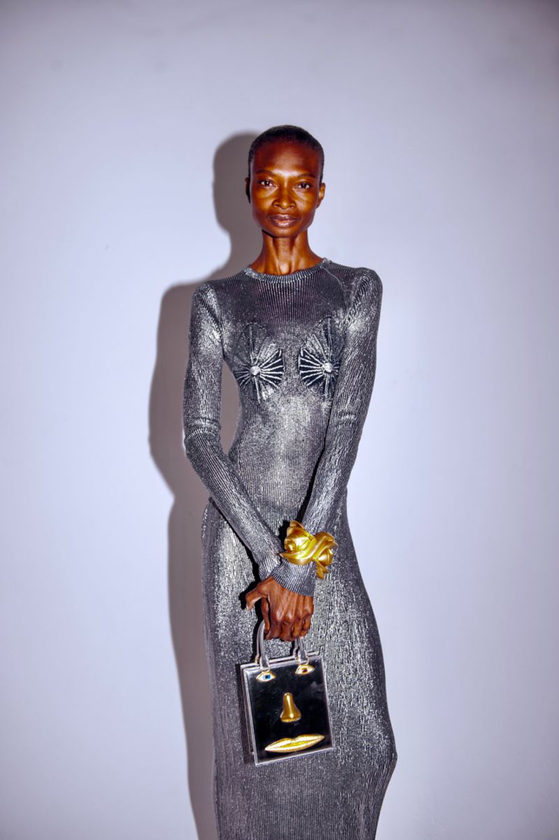 Elegant, slender Black woman poses in long-sleeved silver dress with matching metallic bag and gold cuff in bright studio. 