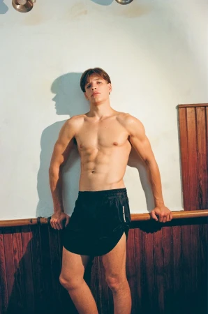 Muscular male athlete poses indoors wearing running shorts, showcasing athletic physique in a fitness environment with natural light.