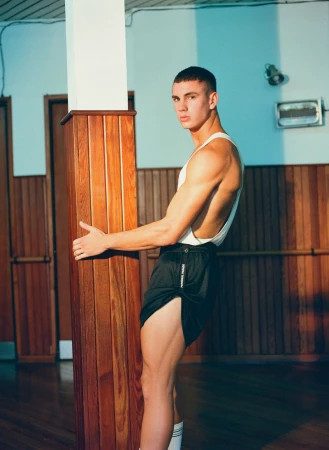 Athletic man in black shorts and white tank poses with a wooden post in an indoor location featuring blue and brown tones.