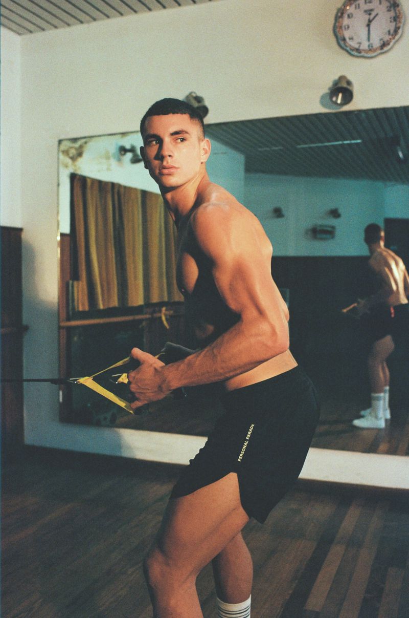 Muscular man in gym shorts and socks doing resistance band workout in a gym with a mirror, clock, and wooden floor.