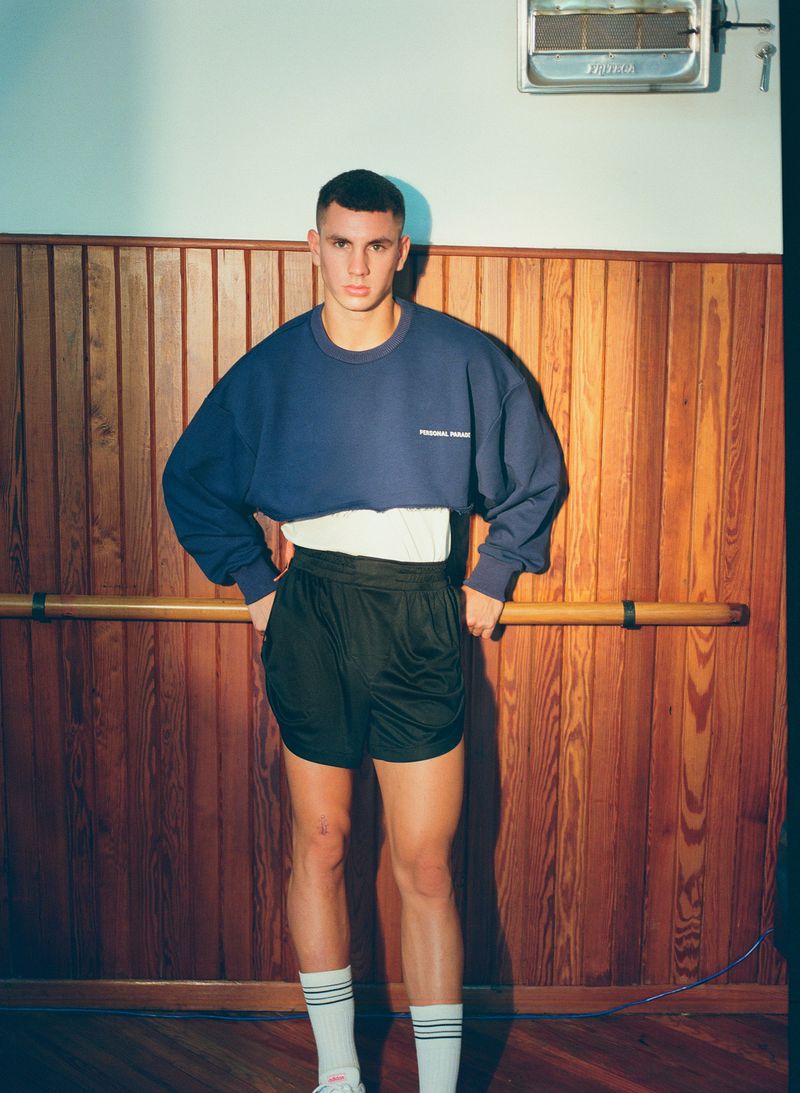 Man in a blue cropped sweatshirt, black shorts, and striped socks poses in a gymnasium with wood paneling and an overhead heater.