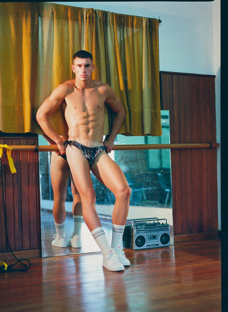 Muscular man in swimwear poses in a dance studio with ballet barre and boombox, showcasing fitness and retro aesthetics in a lifestyle shot.