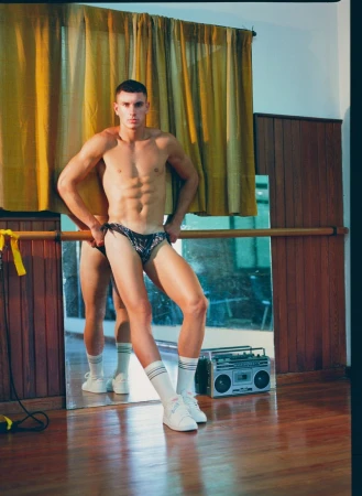 Muscular man in swimwear poses in a dance studio with ballet barre and boombox, showcasing fitness and retro aesthetics in a lifestyle shot.