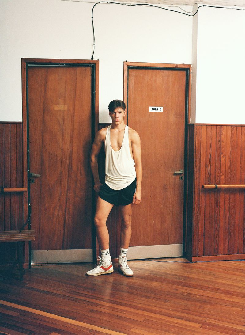 Athletic man in vintage sportswear poses indoors. He wears a white tank top, black shorts, high socks and vintage sneakers in a gym locker room.