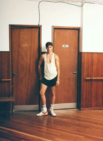 Athletic man in vintage sportswear poses indoors. He wears a white tank top, black shorts, high socks and vintage sneakers in a gym locker room.