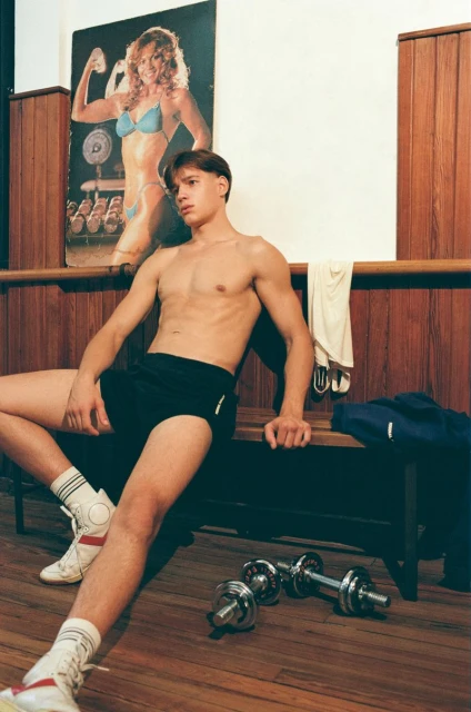 Man in black shorts sitting on a bench indoors next to dumbbells, wearing white socks and athletic shoes, with a poster in background.