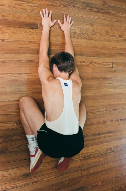 Athletic man stretches out with arms extended upwards, leaning against a wooden floor, wearing a singlet and shorts, from overhead perspective.