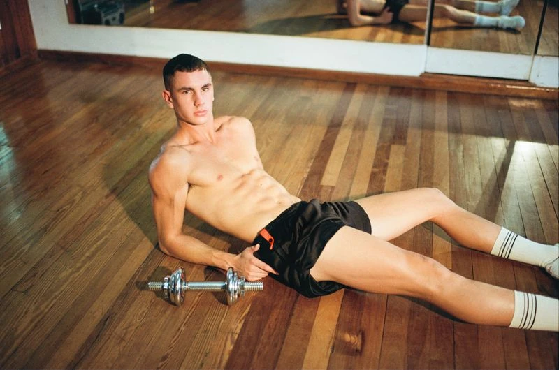Muscular man in shorts and socks lies on a wood floor with a dumbbell, reflecting in a mirror on the wall of an indoor gym.