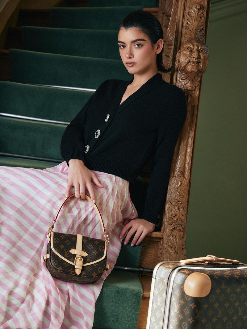 Indoor portrait of a woman sitting on green stairs with a designer purse and a suitcase, wearing a black sweater and pink striped skirt.
