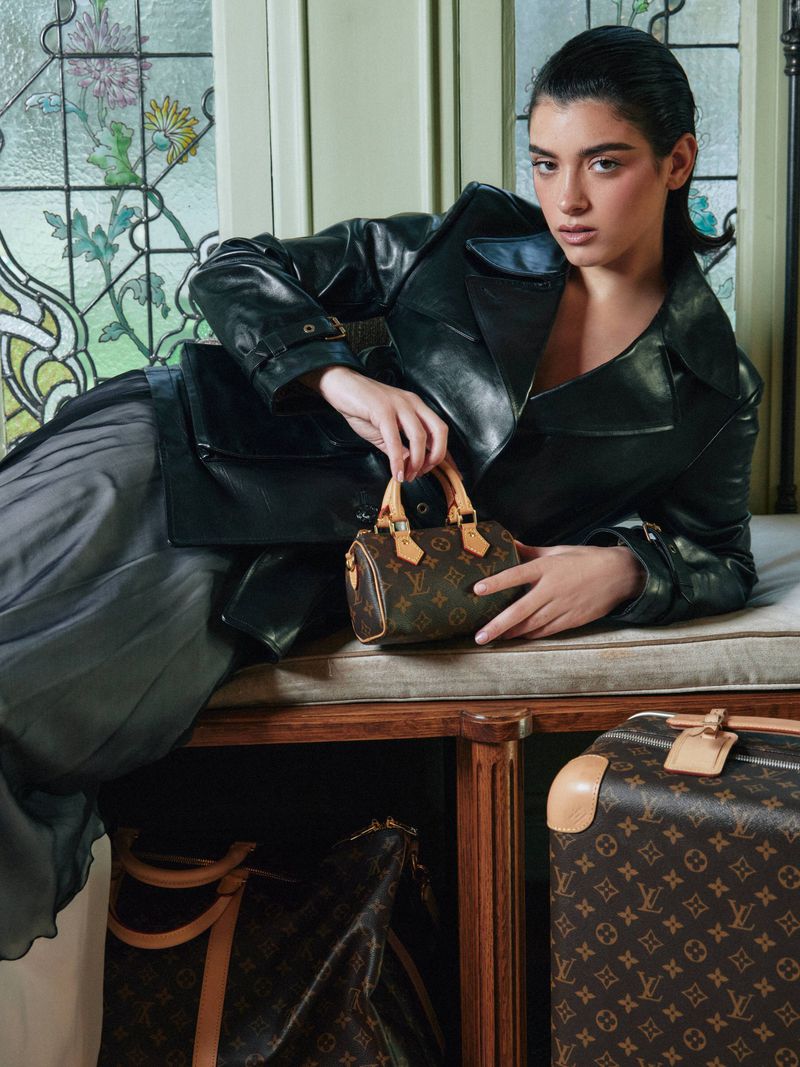 A young woman in a black leather jacket poses with a small Louis Vuitton handbag, next to patterned Louis Vuitton luggage and a stained glass window.