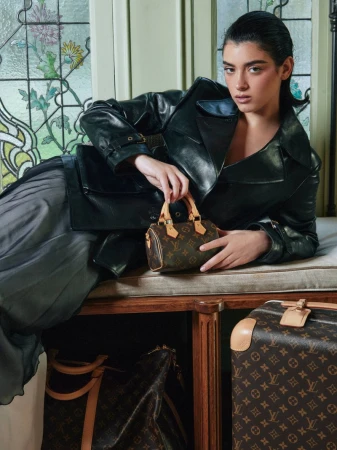 A young woman in a black leather jacket poses with a small Louis Vuitton handbag, next to patterned Louis Vuitton luggage and a stained glass window.