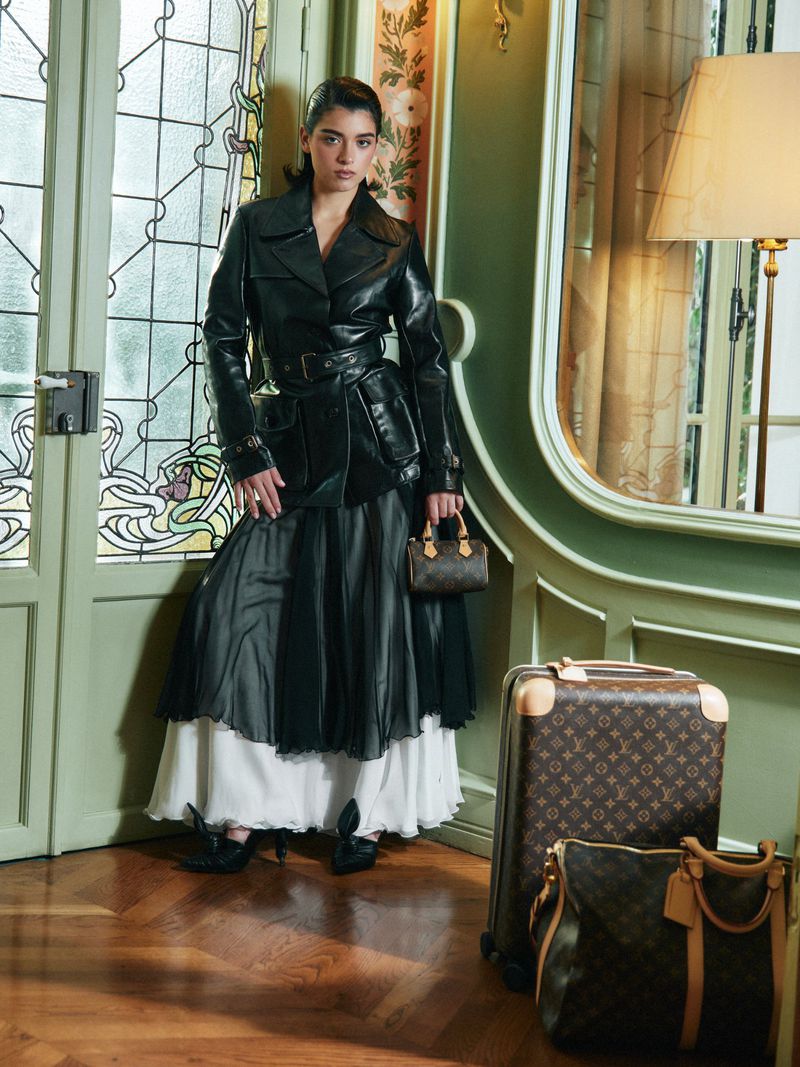 Elegant woman in a black leather trench coat and flowing skirt poses with monogrammed luggage in a vintage-style interior setting.