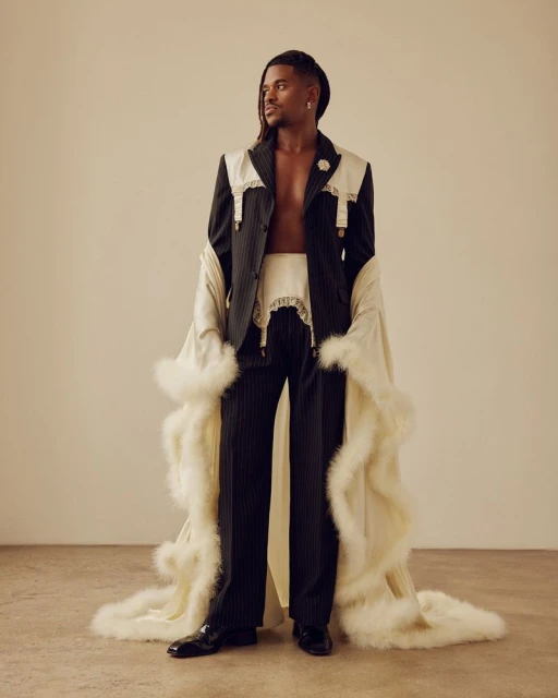 Elegant black man in pinstripe suit with off-white accents, standing in a neutral studio. The man is wearing a robe with feather trim.