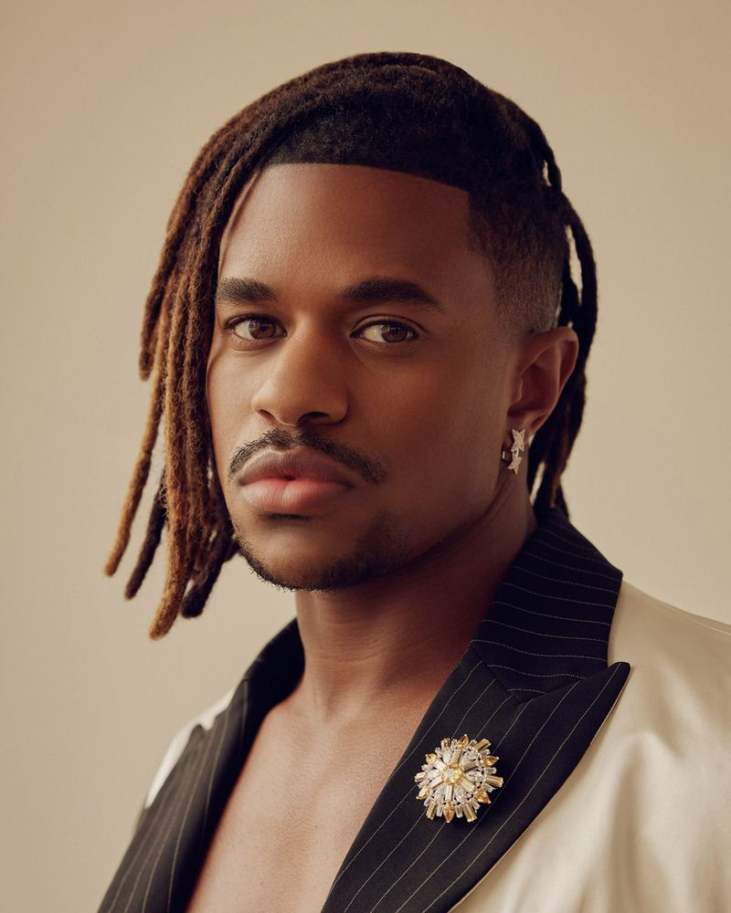 Close-up portrait of a Black male adult with dreadlocks, wearing an off-white blazer with pinstripes and a decorative brooch.