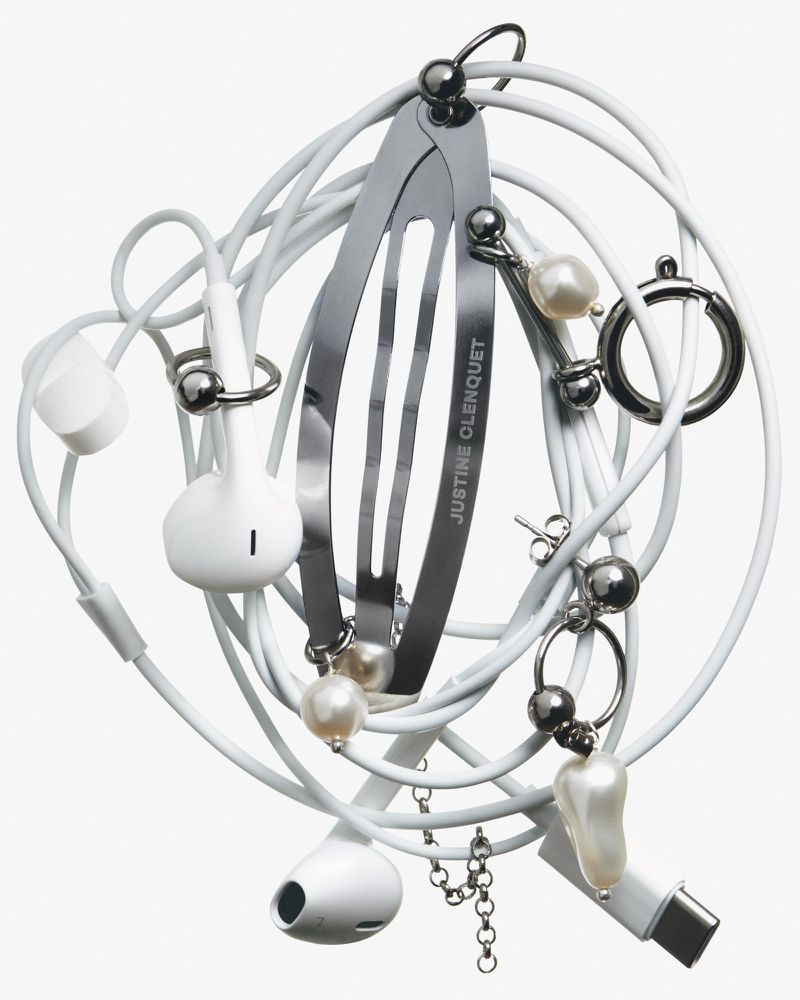 Still life of entangled white Apple earbuds and charging cable, with a metal hair clip and faux pearl jewelry on a white backdrop.