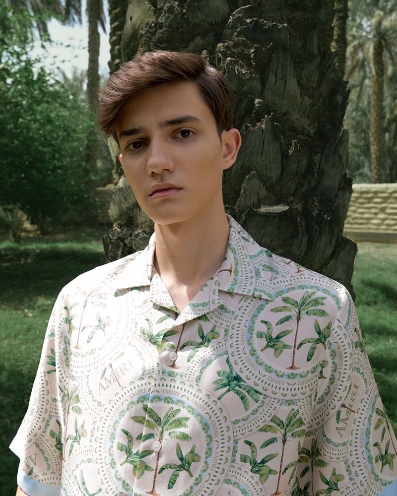 A young man stands outside in patterned button-down shirt, posing in a close up shot with blurred palm tree and vegetation background.