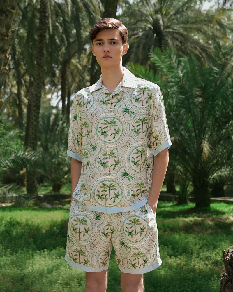 Young man in patterned shirt and shorts posing outdoors against a backdrop of palm trees and green grass on a sunny day.