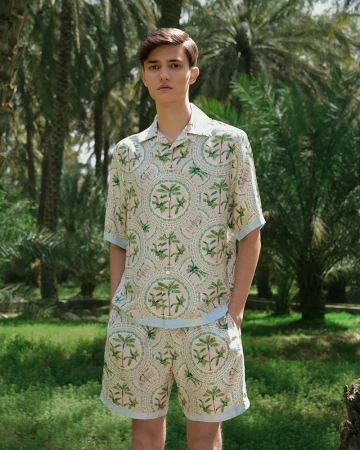 Young man in patterned shirt and shorts posing outdoors against a backdrop of palm trees and green grass on a sunny day.