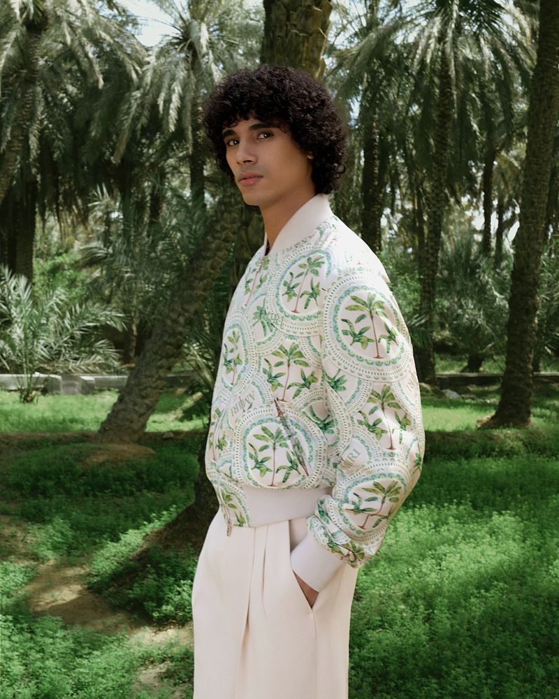 Male model in patterned bomber jacket and tailored pants poses in a palm tree grove. Fashion editorial against a natural backdrop.