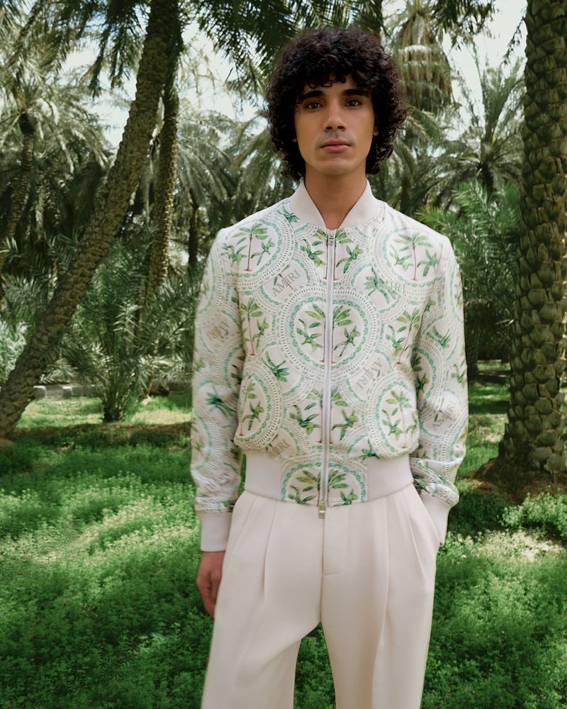 A young adult male wearing a patterned bomber jacket and light-colored pants stands in a grove of palm trees in a medium shot.