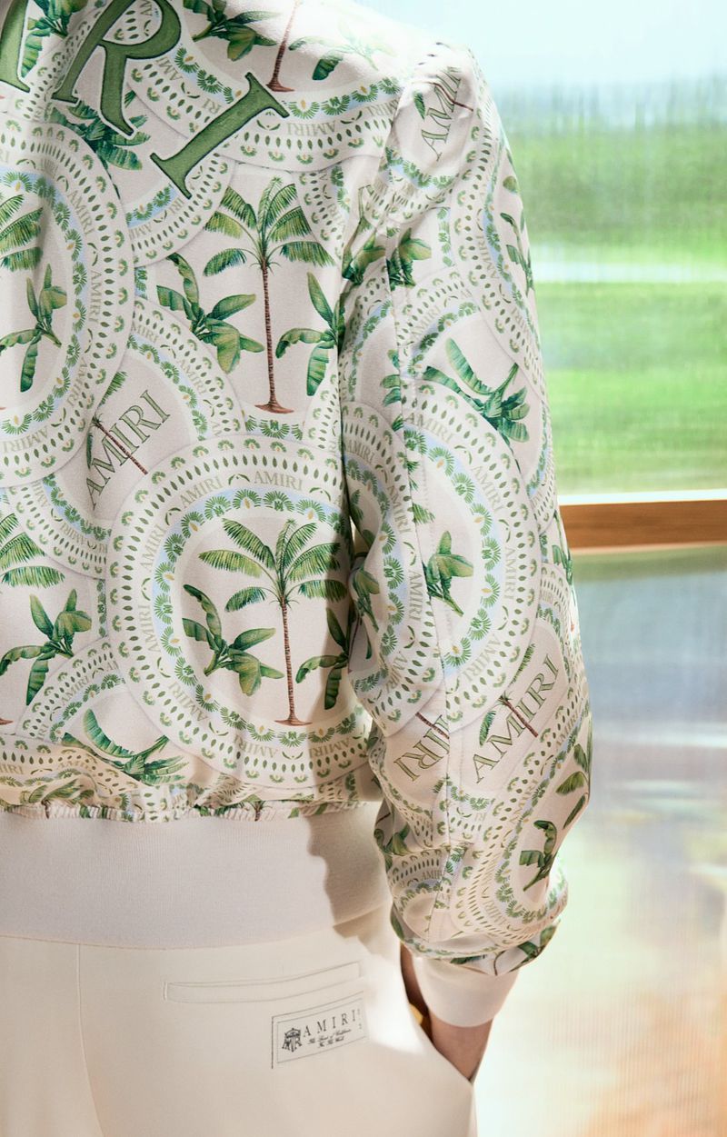 Close-up of a patterned white and green jacket featuring palm tree motifs, paired with white trousers. Fashionable apparel detail.
