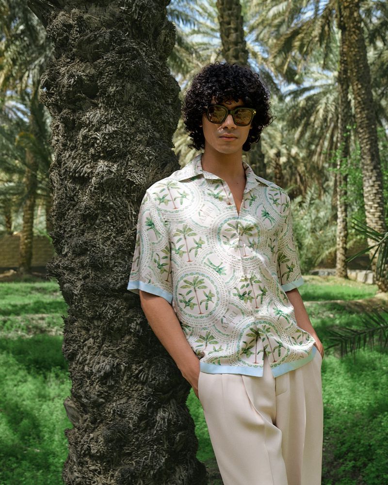 Man in patterned shirt leans against palm tree on sunny day. Outdoor fashion shot in a grove of trees, wearing sunglasses.