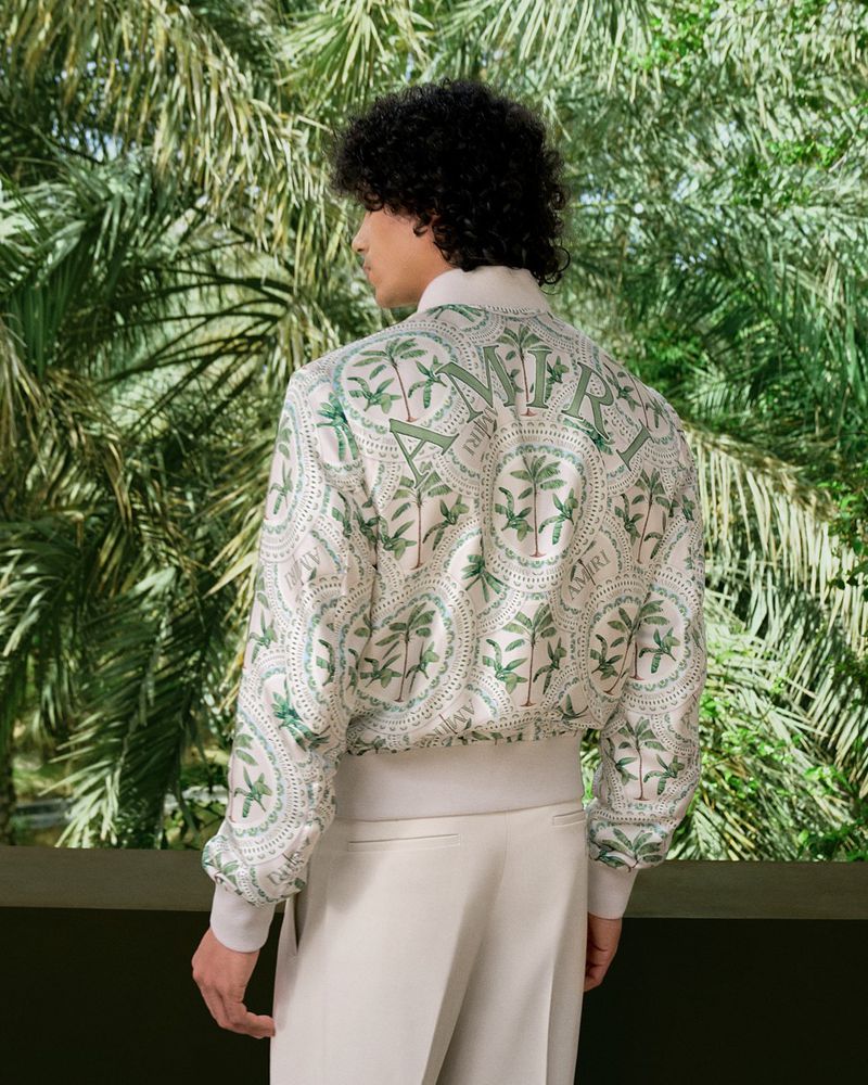 Back view of a male fashion model wearing a patterned silk jacket and cream trousers, posing against a backdrop of palm trees.