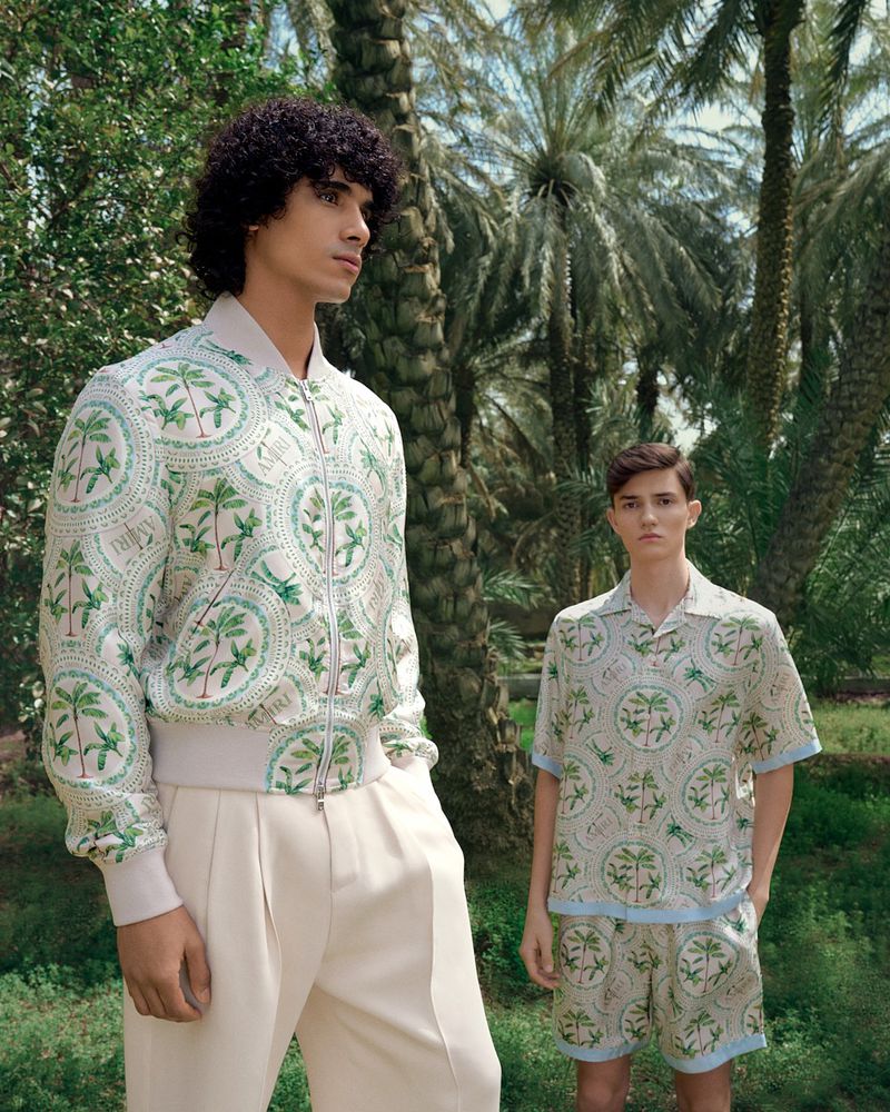 Two young men pose outdoors in matching patterned outfits. The man in front wears a bomber jacket with slacks; the other wears a shirt and shorts.