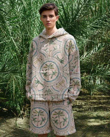 Man poses outdoors wearing a cream-colored patterned hoodie and shorts with palm tree design, set against a lush green foliage backdrop.