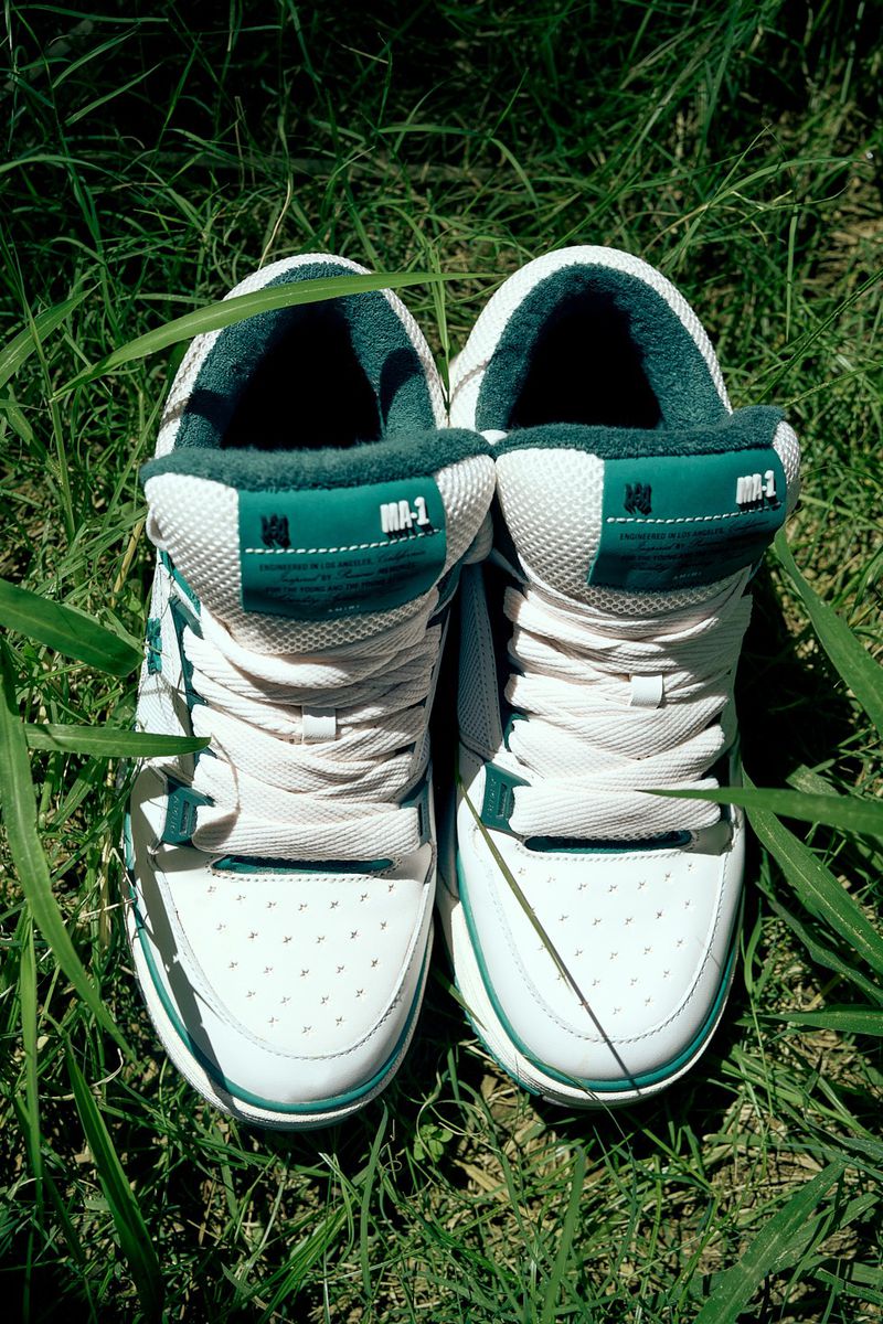 Pair of white and green athletic shoes sit in green grass, laces tied, soles visible, in natural light. Outdoor setting.