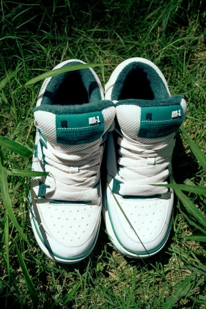 Pair of white and green athletic shoes sit in green grass, laces tied, soles visible, in natural light. Outdoor setting.