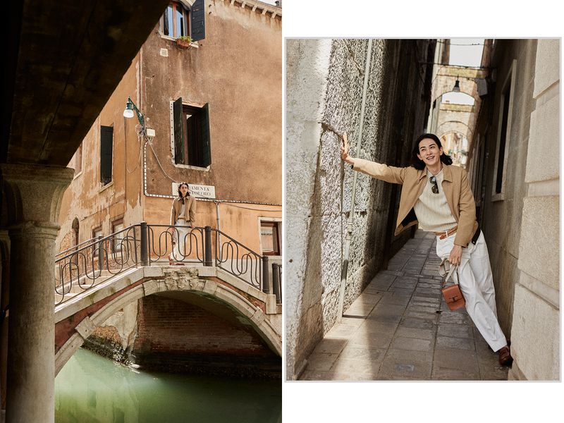 A woman in a tan jacket and white pants stands on a bridge in Venice, Italy, with buildings and a canal visible in the background.