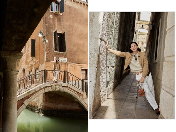 A woman in a tan jacket and white pants stands on a bridge in Venice, Italy, with buildings and a canal visible in the background.