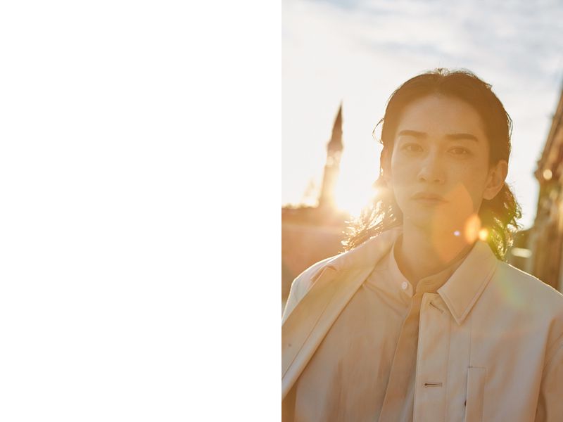Portrait of a young adult male outdoors in warm sunlight wearing a jacket and shirt. Historic building in background; fashion shot.