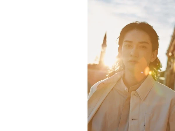 Portrait of a young adult male outdoors in warm sunlight wearing a jacket and shirt. Historic building in background; fashion shot.