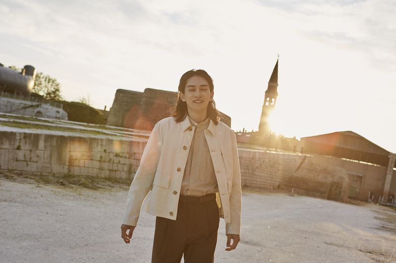 Adult male fashion model in a beige jacket and brown pants poses outdoors with historic buildings and warm, late-afternoon sun.