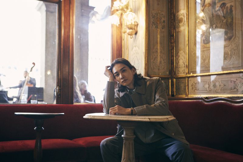 Adult male with long hair wearing a coat, sitting at a table in a cafe setting with a red bench seat. Natural light.
