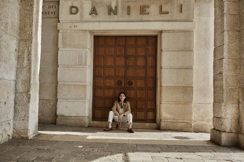 Young adult woman sitting in front of a grand wooden door in a stone archway in an urban environment with warm, natural lighting.