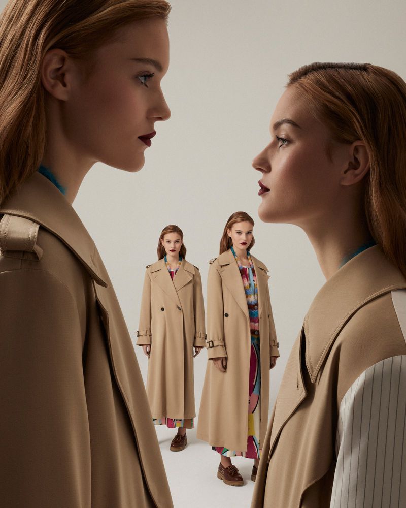 Fashion shot featuring two red-haired women in tan trench coats against a pale background, striking a pose that accentuates the clothing's style.
